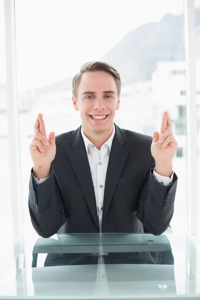 Smiling businessman with fingers crossed at office desk — Stock Photo, Image