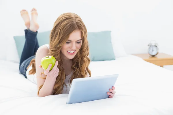 按住一个苹果并在床上使用 tablet pc 的休闲红头发的女孩 — 图库照片