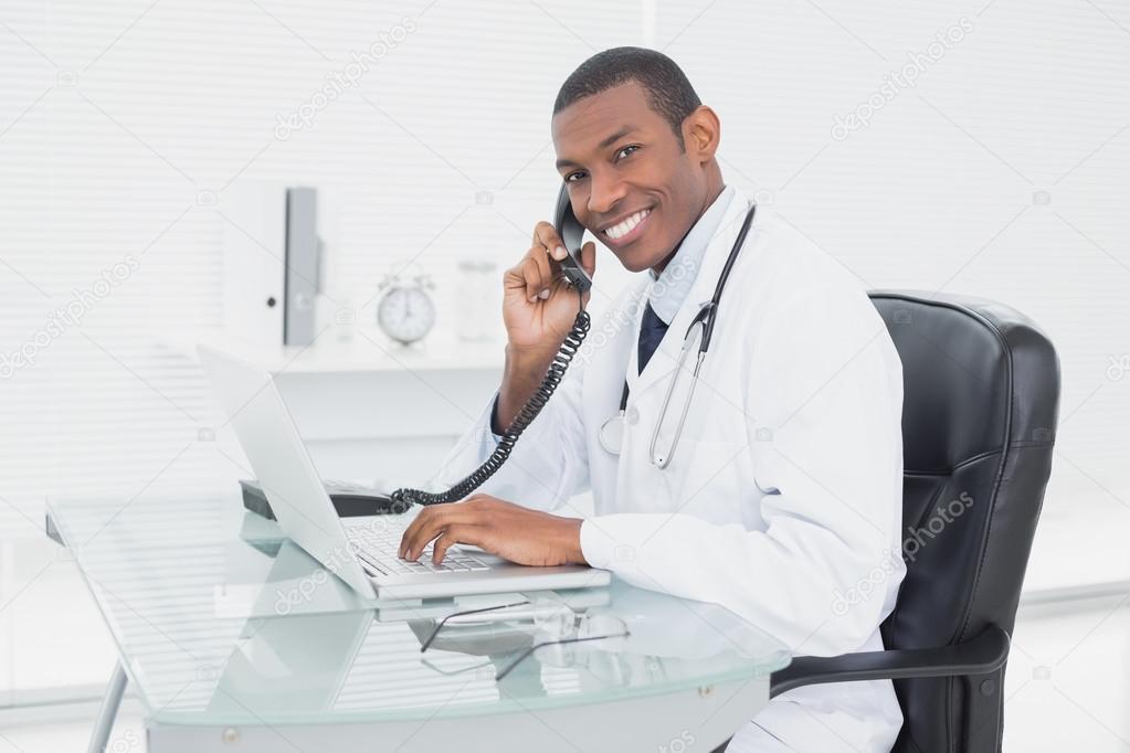 Smiling doctor using phone and laptop at medical office