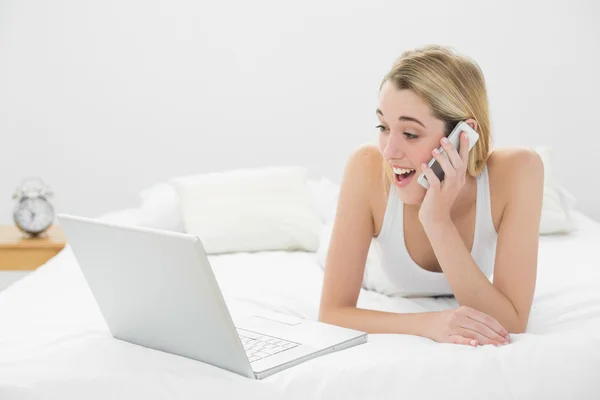 Mujer encantadora asombrada llamando con su teléfono inteligente mirando su computadora portátil —  Fotos de Stock