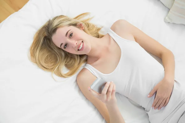 Gioiosa donna bionda che tiene il suo smartphone sdraiato sul letto — Foto Stock