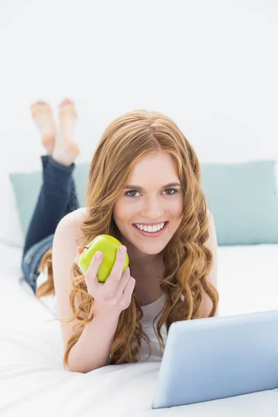 Casual roodharige meisje tablet pc gebruikt terwijl een appel in bed — Stockfoto