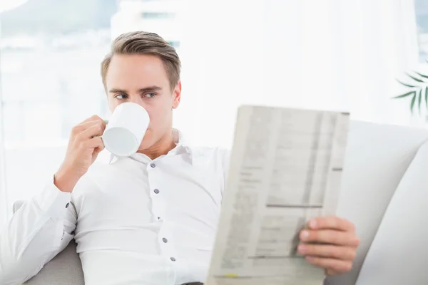 Relajado hombre bebiendo café mientras lee el periódico en el sofá —  Fotos de Stock