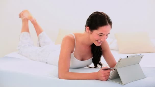 Приваблива брюнетка використовує свій цифровий планшет, щоб купити онлайн лежачи на ліжку — стокове відео