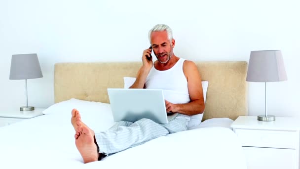 Hombre sonriente usando el ordenador portátil mientras está en el teléfono — Vídeos de Stock