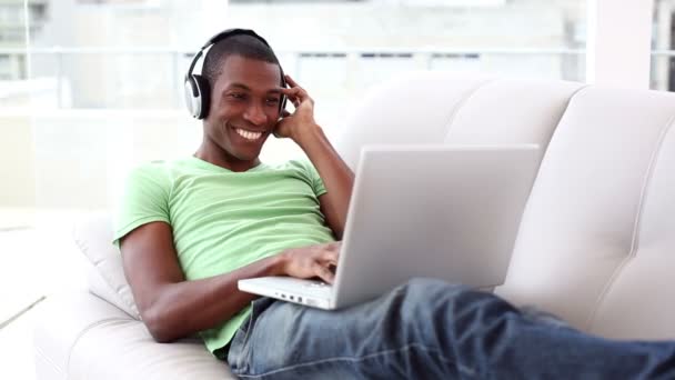 Улыбающийся мужчина лежит на диване и слушает музыку с помощью ноутбука — стоковое видео