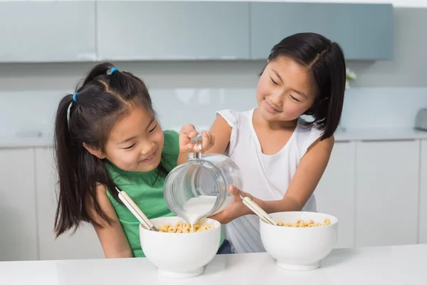 Two happy young girls pouring milk in bowl in kitchen — Stock Photo, Image