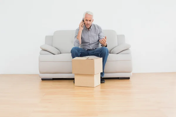 Uomo maturo con cellpone sul divano con scatole in una casa — Foto Stock