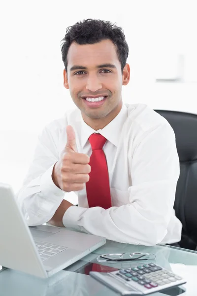 Uomo d'affari sorridente con computer portatile gesticolando pollici in alto in ufficio — Foto Stock