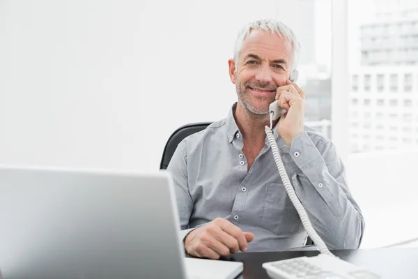Smiling businessman on call in front of laptop at office desk — Stock Photo, Image