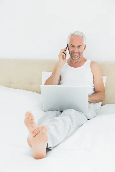 Casual χαμογελώντας ώριμος άνδρας, χρησιμοποιώντας το κινητό και το laptop στο κρεβάτι — Φωτογραφία Αρχείου