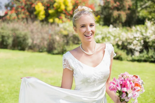 Beautiful blonde bride holding bouquet and her dress smiling at camera — Stock Photo, Image