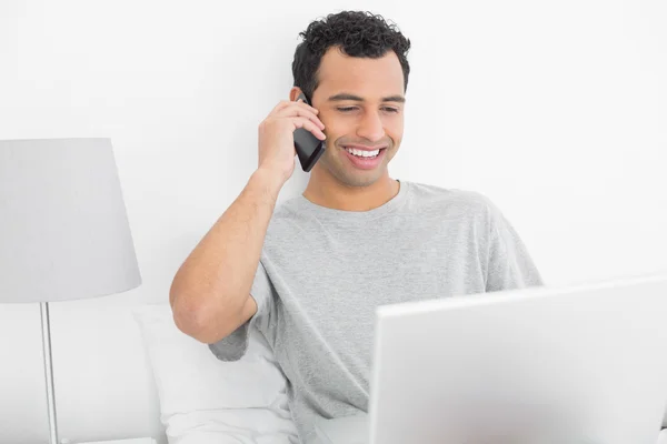 Homme souriant occasionnel utilisant un téléphone portable et un ordinateur portable au lit — Photo