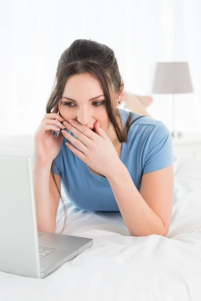 Mulher chocada usando telefone celular e laptop na cama — Fotografia de Stock