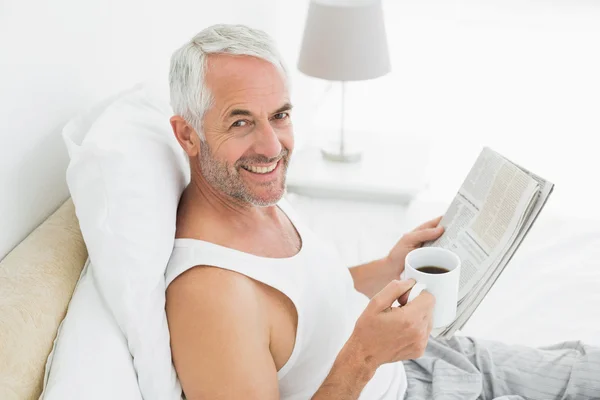 成熟的男人微笑着与杯咖啡和报纸躺在床上 — 图库照片