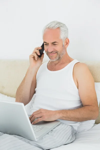 Olgun erkek yatakta kullanarak cep telefonu ve laptop — Stok fotoğraf