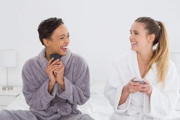 Cheerful friends in bathrobes text messaging on bed — Stock Photo, Image