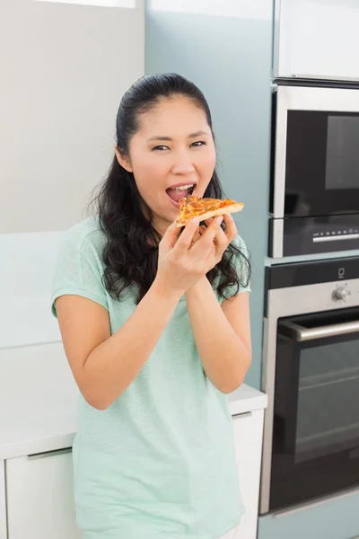 Smiling young woman eating a slice of pizza in kitchen — Stock Photo, Image