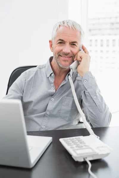Businessman using telephone in front of laptop at office desk — Stock Photo, Image