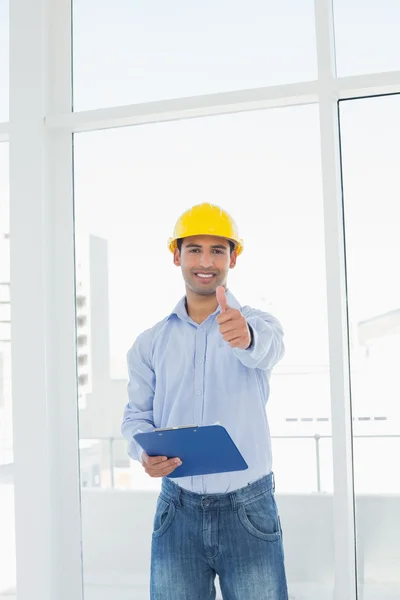 Architect in hard hat with clipboard gesturing thumbs up in office — Stock Photo, Image