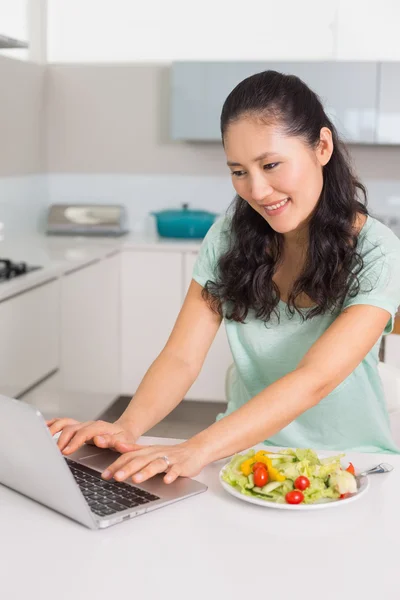 Mujer sonriente usando portátil mientras tiene ensalada en la cocina —  Fotos de Stock