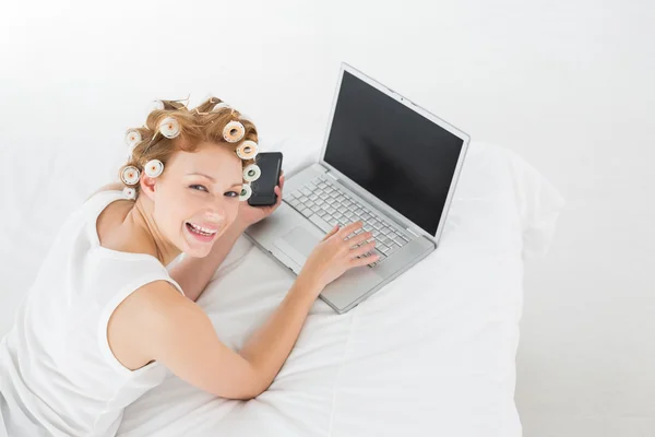 Donna allegra in bigodini per capelli utilizzando il computer portatile a letto — Foto Stock