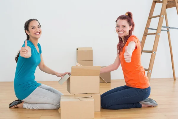 Female friends gesturing thumbs up with boxes in a new house — Stock Photo, Image