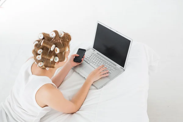 Woman in hair curlers with cellphone using laptop in bed — Stock Photo, Image