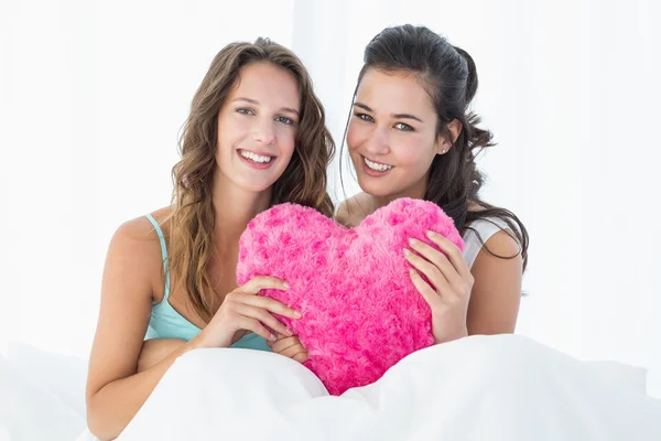 Sonrientes amigas con almohada en forma de corazón en la cama —  Fotos de Stock