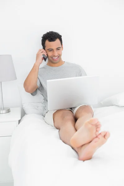 Hombre sonriente casual usando teléfono celular y portátil en la cama —  Fotos de Stock