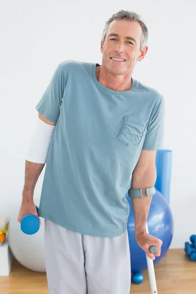 Smiling mature man with crutch and dumbbell — Stock Photo, Image