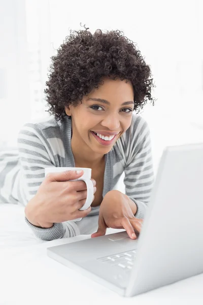 Lachende vrouw met koffie beker met behulp van laptop in bed — Stockfoto