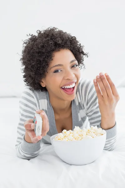 Veselá žena s dálkovým ovládáním a mísa popcornu v posteli — Stock fotografie