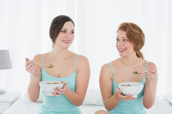 Leende kvinnliga vänner med salladsskålar sitter på sängen — Stockfoto