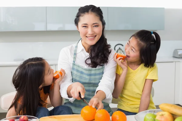 Woman with happy two daughters cutting fruit in kitchen — Stock Photo, Image