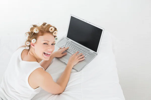 Cheerful woman in hair curlers using laptop in bed — Stock Photo, Image