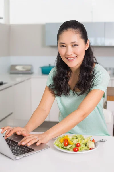 Gülümseyen kadın mutfakta salatası yerken dizüstü kullanarak — Stok fotoğraf