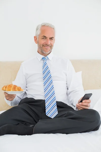 Geschäftsmann schreibt SMS, während er Croissant im Bett hält — Stockfoto