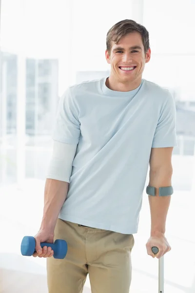 Smiling young man with crutch and dumbbell — Stock Photo, Image