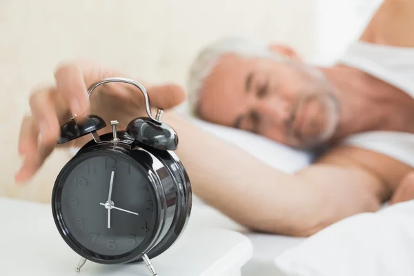 中高年の男性がベッドの中でアラーム時計手を拡張します。 — ストック写真