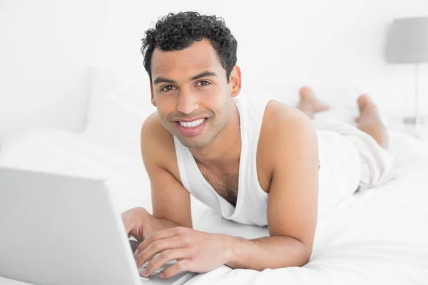 Χαμογελώντας casual νεαρός χρησιμοποιώντας φορητό υπολογιστή στο κρεβάτι — Φωτογραφία Αρχείου