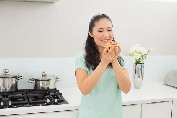 Sorrindo jovem mulher comendo uma fatia de pizza na cozinha — Fotografia de Stock