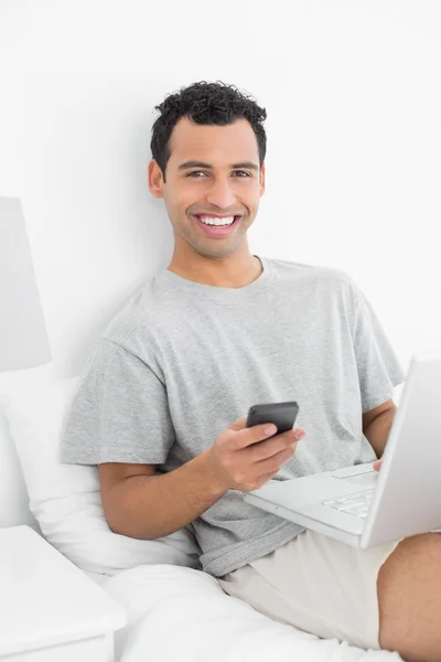 Hombre sonriente casual con mensajería de texto portátil en la cama —  Fotos de Stock