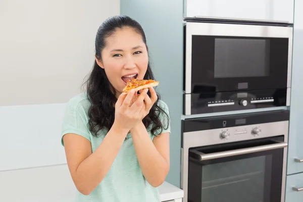 Smiling young woman eating a slice of pizza in kitchen — Stock Photo, Image