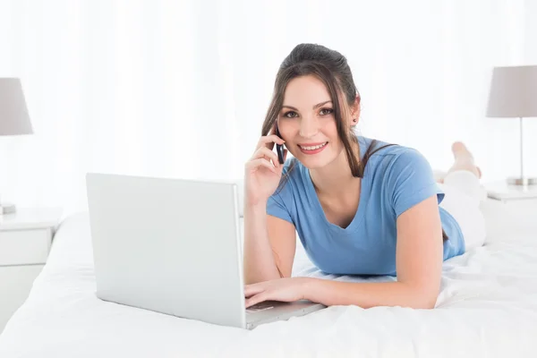 Smiling woman using mobile phone and laptop in bed — Stock Photo, Image