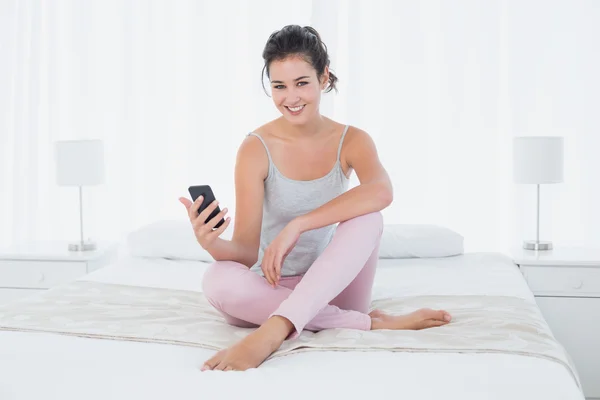 Lachende vrouw die zit met mobiele telefoon in bed — Stockfoto