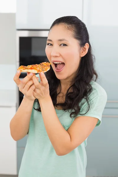 Sorrindo jovem mulher comendo uma fatia de pizza na cozinha — Fotografia de Stock