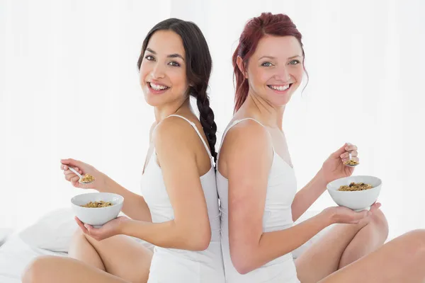 Side view of smiling friends with bowls sitting on bed — Stock Photo, Image