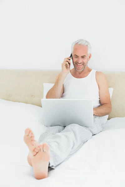 Sonriendo hombre maduro utilizando el teléfono celular y el ordenador portátil en la cama —  Fotos de Stock