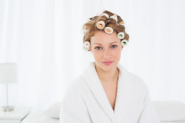 Beautiful woman in bathrobe and hair curlers sitting on bed — Stock Photo, Image